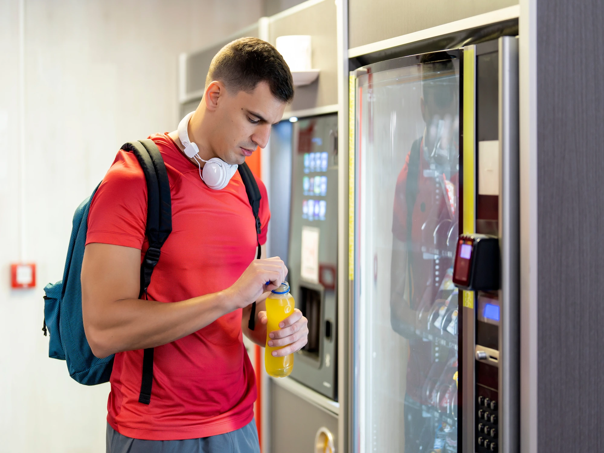 Young male athlete with backpack and headphones opening drink he has purchased from vending machine. 